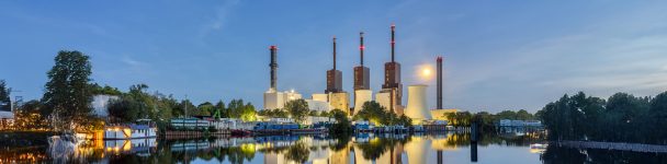 Panoramaaufnahme eines modernen Kraftwerks mit mehreren Kühltürmen und Schornsteinen am Wasser, bei Abenddämmerung.