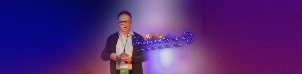 Vortragender Mann mit Mikrofon und Konferenzunterlagen auf der Bühne vor dunklem, buntem Hintergrund mit leuchtendem Schriftzug „NetNight Inspiration 23“ – Szene eines IT-Events oder Fachvortrags.