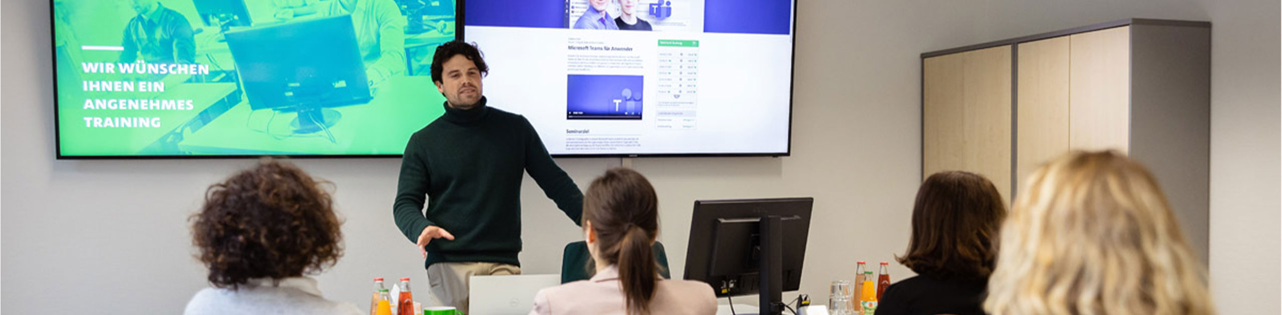 Ein IT-Dozent erklärt Inhalte vor Teilnehmer:innen in einem modernen Seminarraum. Auf dem Bildschirm sind Schulungsinhalte zu Microsoft Teams sowie der Text „Wir wünschen Ihnen ein angenehmes Training“ zu sehen.